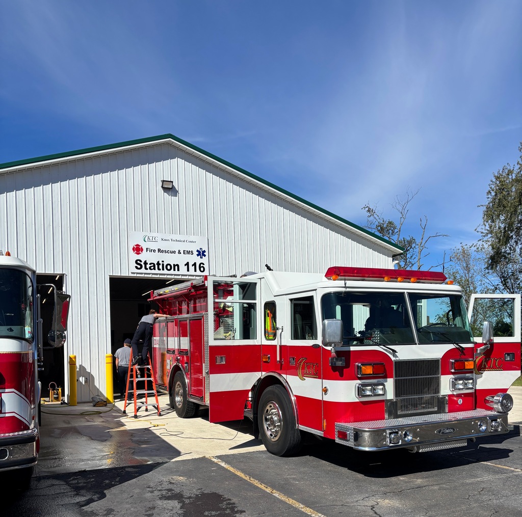 A red and white fire engine parked outside a white building labeled "KTC Fire Rescue & EMS Station 116" while firefighters conduct maintenance.