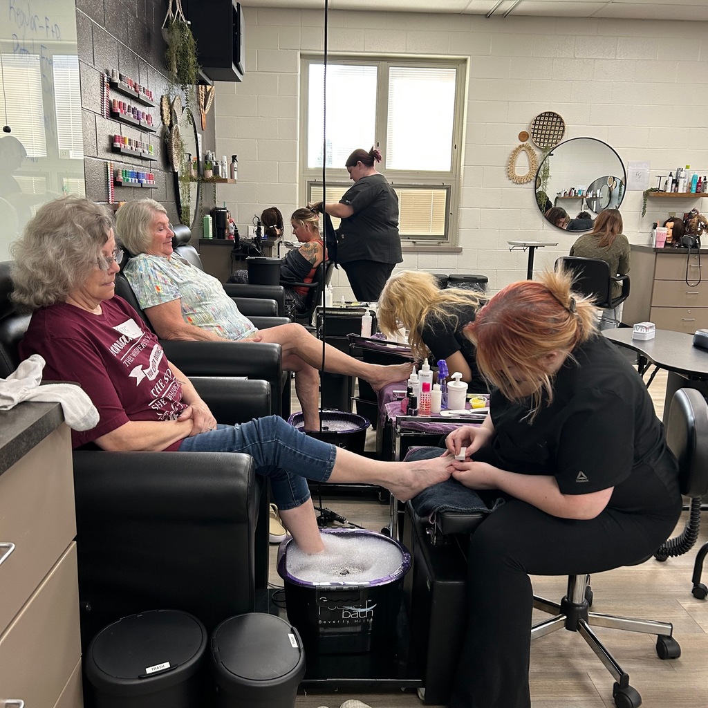 Cosmetology students in black scrubs provide pedicure and hair services to clients in a well-equipped salon classroom