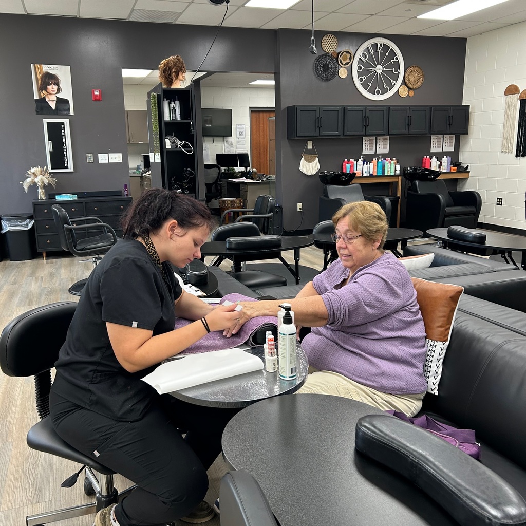 A cosmetology student in black scrubs giving a manicure to a client in a purple sweater seated at a salon station.