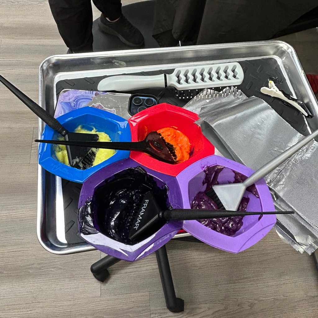 An overhead view of a metal salon tray holding colorful bowls of hair dye, color brushes, foil sheets, and hair tools.