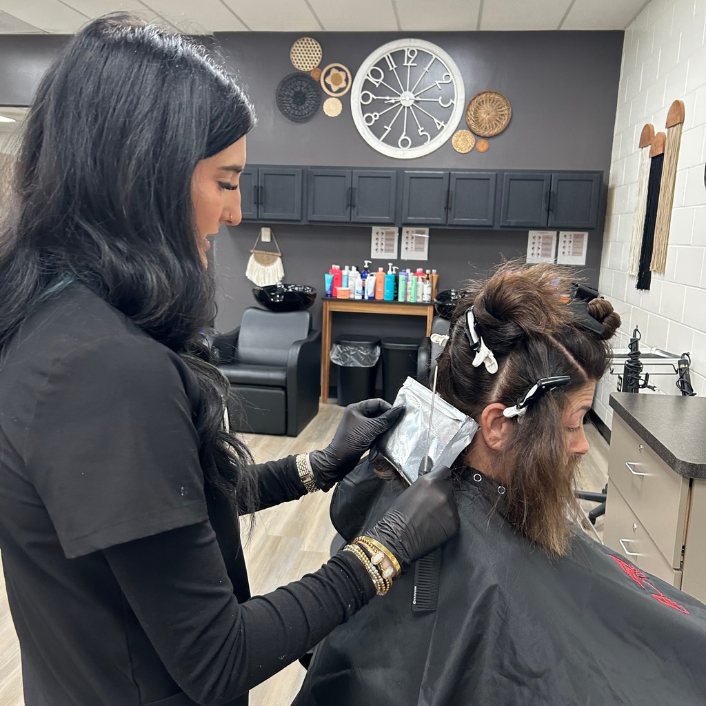 A cosmetology student in black scrubs and black gloves applying hair foils to a client's hair in a salon classroom.
