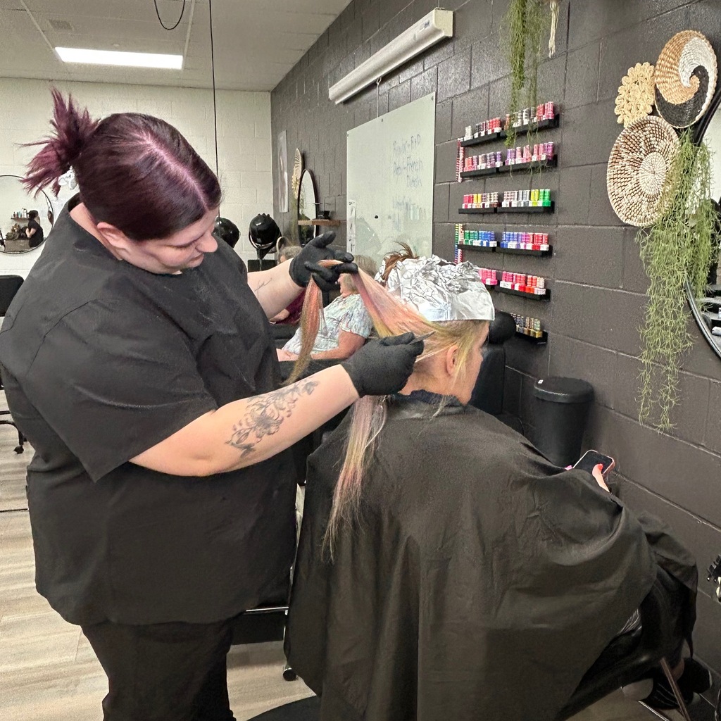 A cosmetology student in black scrubs and black gloves applying hair foils to a client's hair in a salon classroom