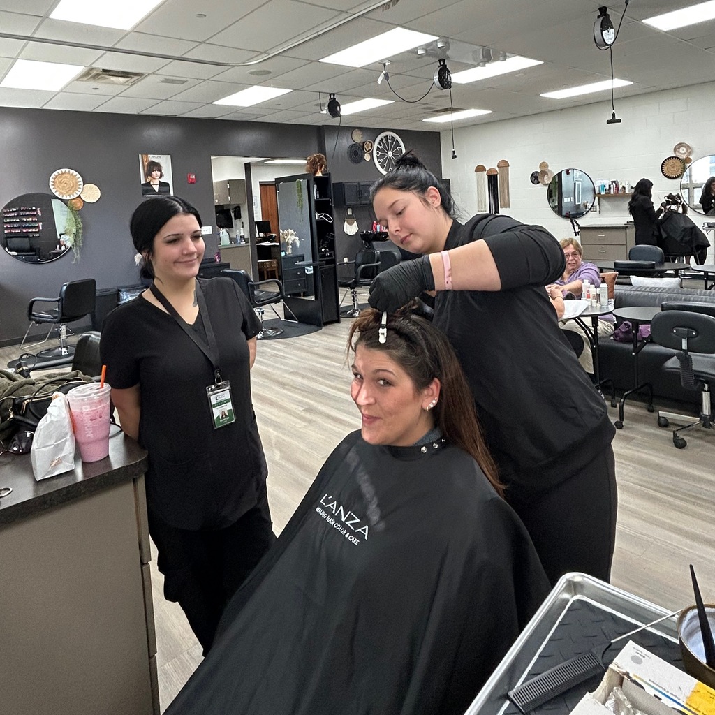In a cosmetology lab, one student in black scrubs trims a client's hair with scissors while another student watches and smiles