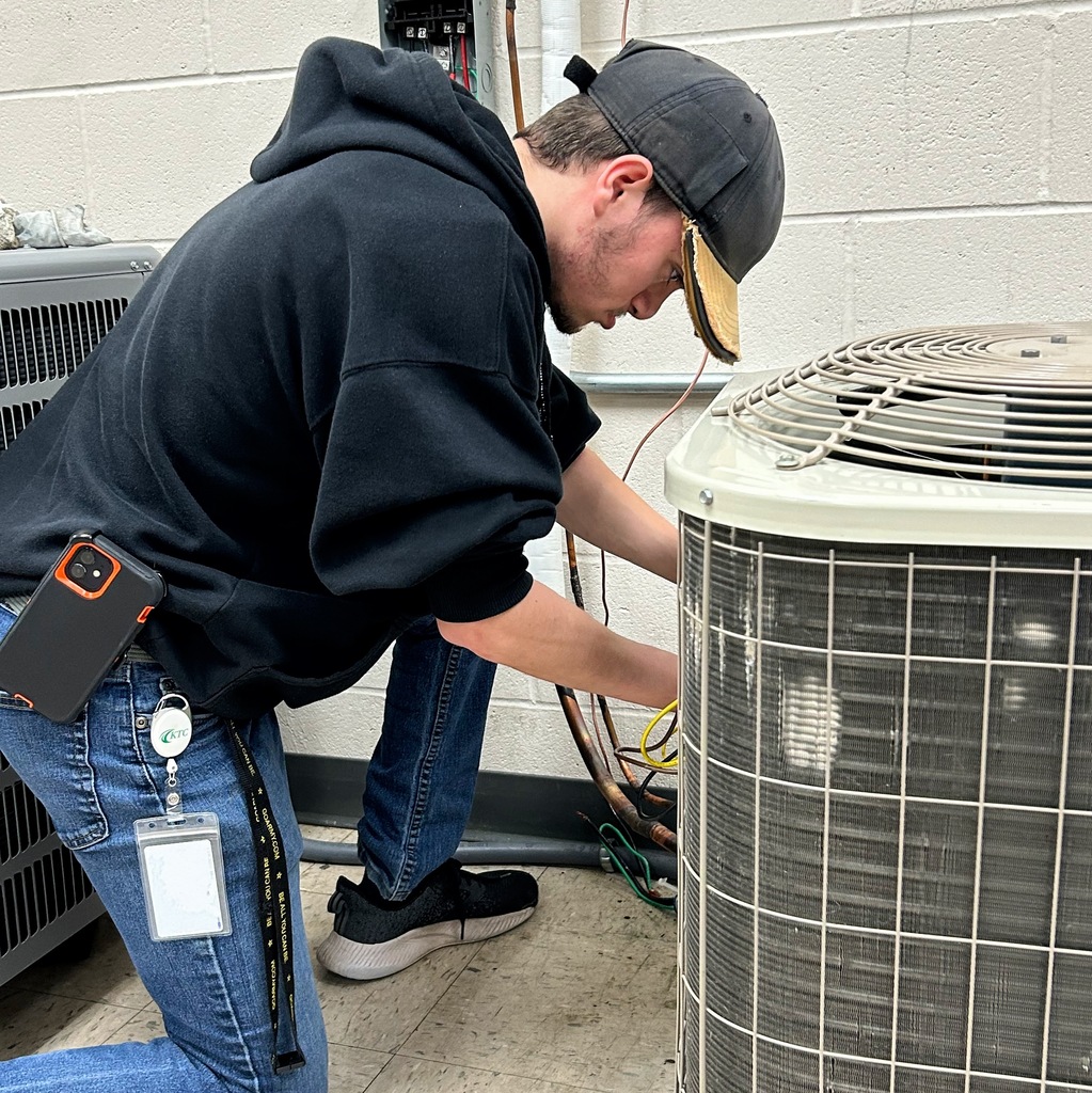 A KTC HVACR student kneels while servicing the wiring and copper lines on an air conditioning unit.