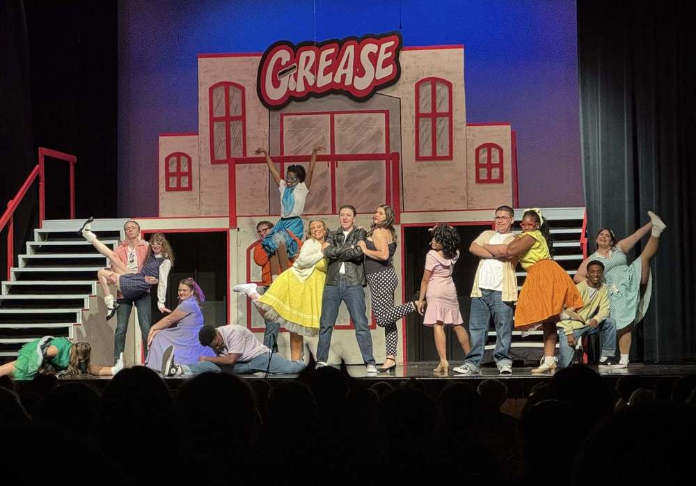 Cast posing on the stage in front of a set with windows and stairs in 50s outfits