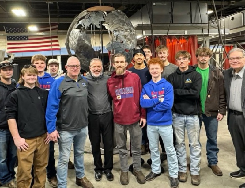 group of student welders standing in welding bay in front of metal globe sculpture with american flag in background