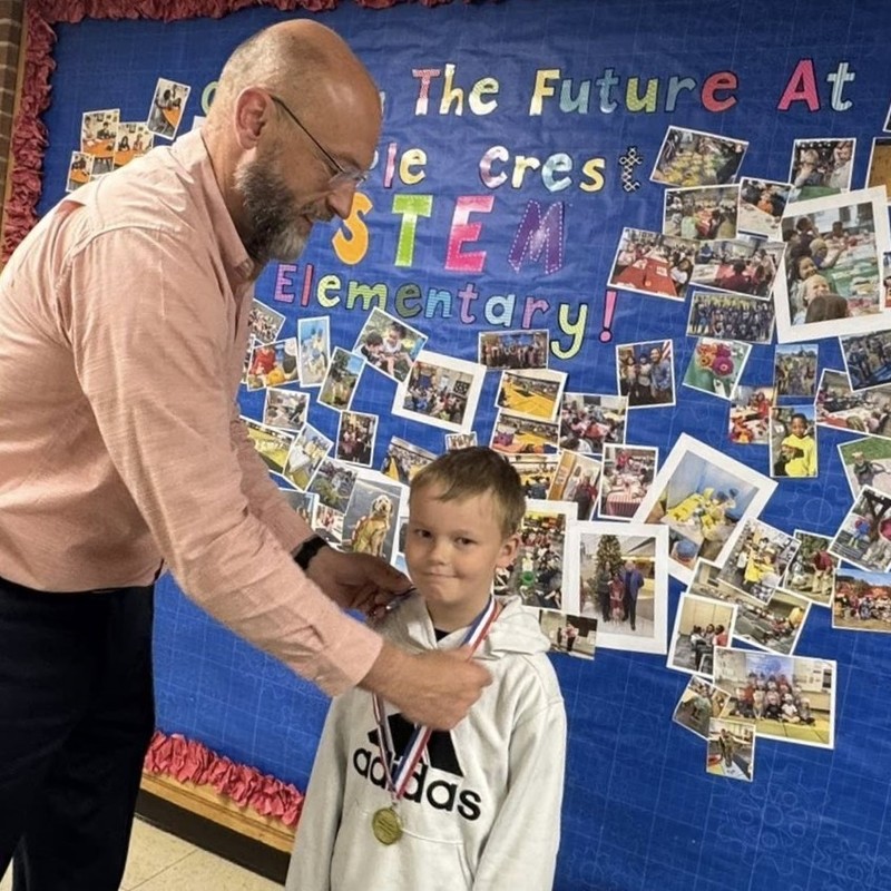 man wearing glasses and smiling placing mathematics award medallion around elementary male student neck