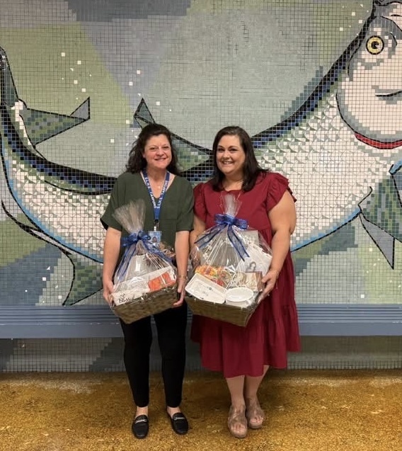 Two smiling women stand indoors holding gift baskets wrapped in clear plastic with blue bows. One wears a green blouse and black pants; the other wears a red dress. Behind them is a mosaic mural of two large silver fish on a blue‑green background.