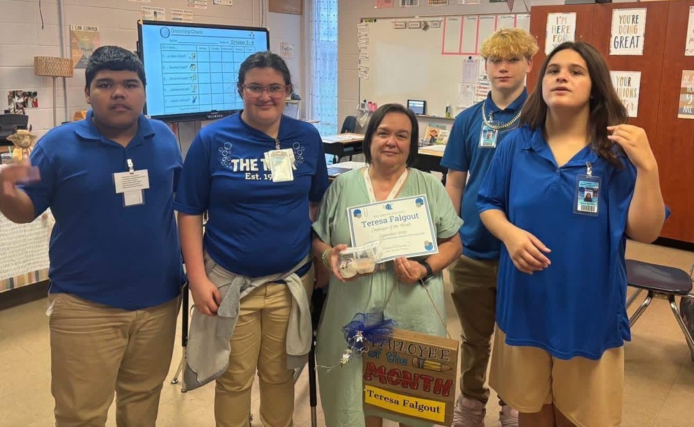 A small group of students stands with a staff member in a classroom. The staff member holds a certificate and an “Employee of the Month” sign while students pose around them. Classroom posters, a whiteboard, and cabinets are visible in the background.