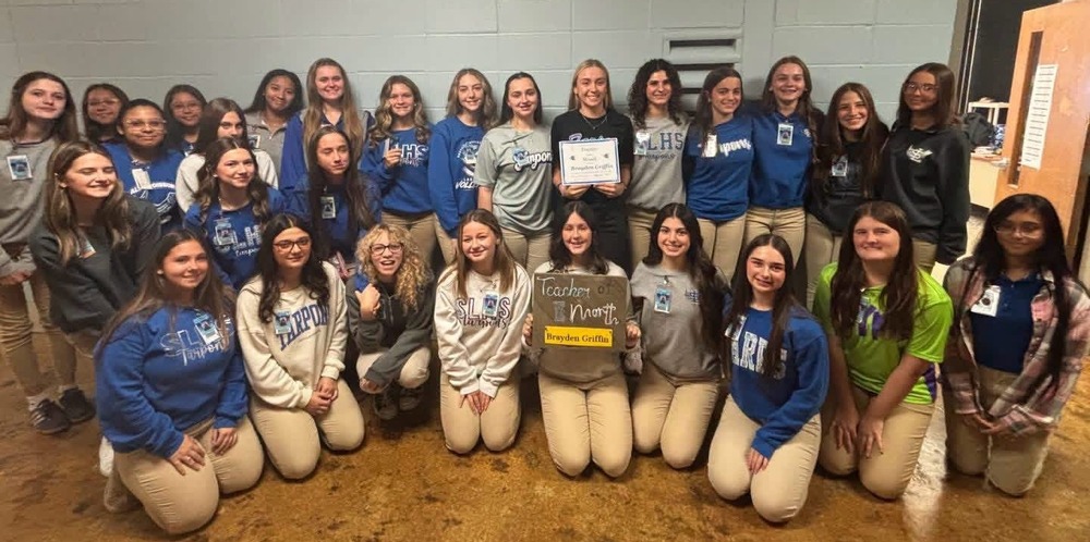 Group photo of approximately 30 students in school uniforms posing indoors with certificates and a “Teacher of the Month” sign. The students are arranged in rows against a light-colored wall in a school hallway or classroom setting.