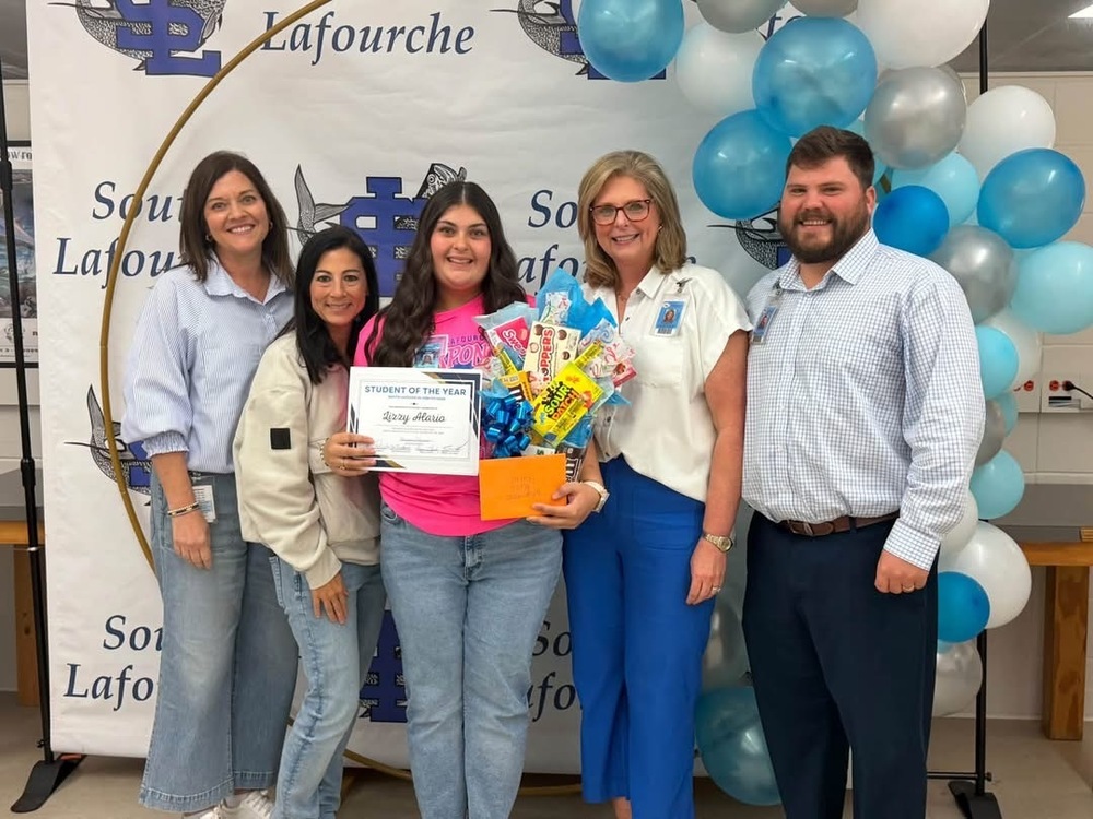 Five people stand smiling in front of a “South Lafourche” backdrop decorated with blue, silver, and white balloons. The person in the center holds a certificate reading “Student of the Year – Lizzy Alario" and a colorful candy bouquet with a blue ribbon. The group appears to be celebrating the student’s achievement.