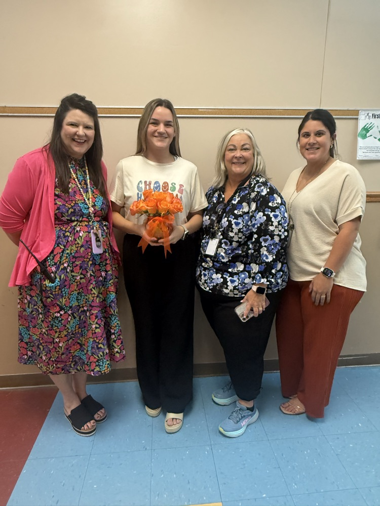 Four adults posing indoors against a beige wall with a blue and red tiled floor. One person holds a small orange bouquet of flowers. The group wears colorful, casual clothing, and the setting appears to be a school or office environment.