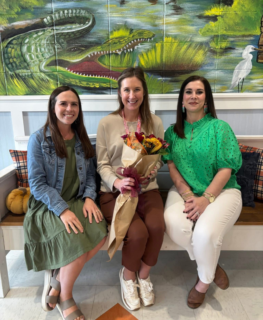 Three individuals are seated on a bench with plaid cushions. The person in the center holds a bouquet of flowers wrapped in brown paper and tied with a ribbon. Behind them is a vibrant mural featuring an alligator, water, greenery, and birds. A small pumpkin is placed on the left side of the bench. The setting appears to be indoors, possibly in a school or community space.