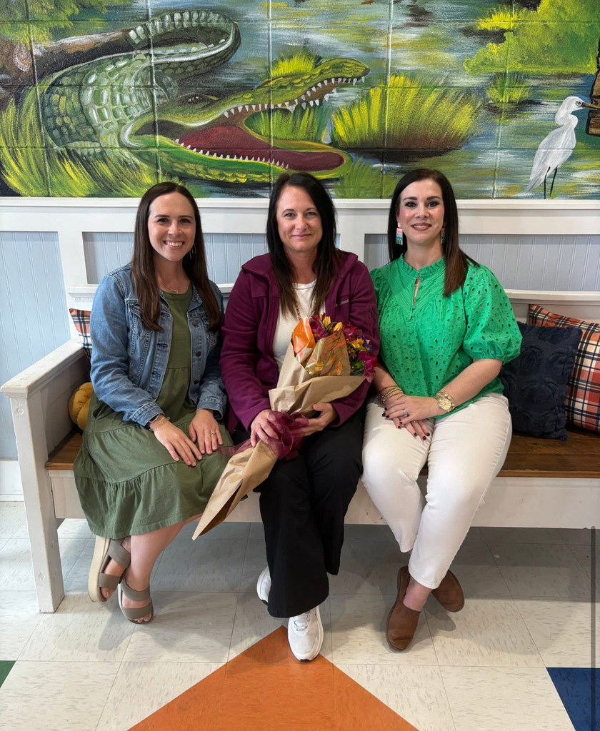 Three individuals are seated on a wooden bench indoors, likely in a school or community center. The person in the center holds a bouquet of flowers wrapped in brown paper. Behind them is a colorful mural depicting an alligator and a heron in a natural setting. The scene suggests a moment of celebration or recognition.