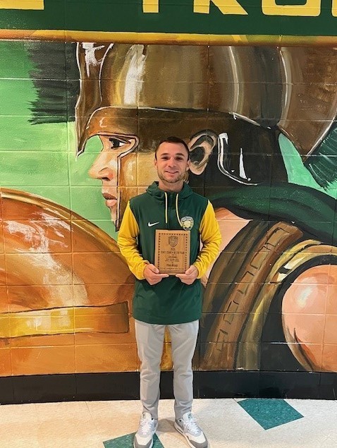 CLHS girls soccer coach Ryan Breaux holding the State Coach of the Year Award in front of a wall with a painted Trojan.