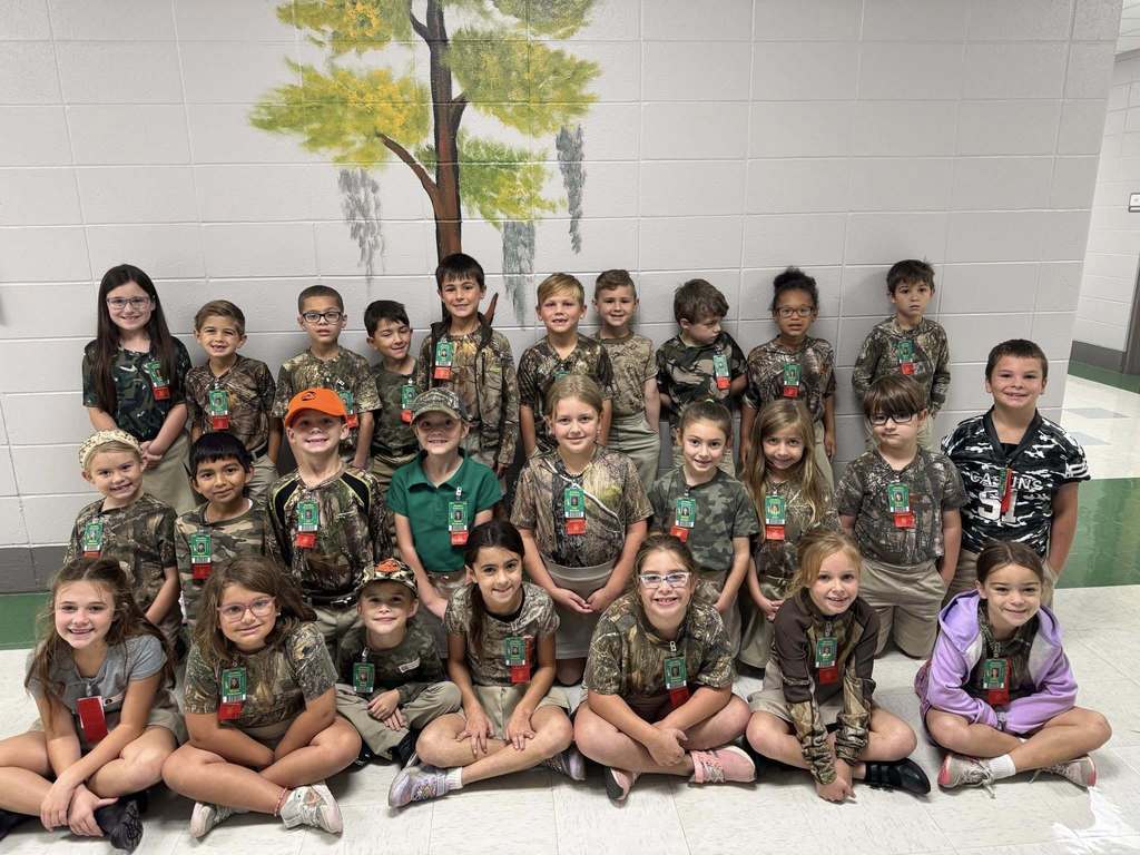 Group of children wearing bright, colorful shirts and khaki bottoms posing in front of a backdrop that says “RED RIBBON” with a large glittery red ribbon, celebrating Red Ribbon Week.
