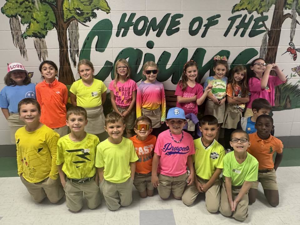 Group of children wearing bright, colorful shirts and khaki bottoms posing in front of a backdrop that says “RED RIBBON” with a large glittery red ribbon, celebrating Red Ribbon Week.