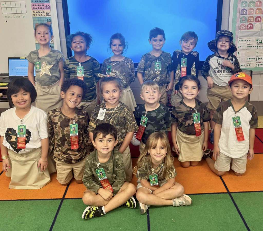 Group of children wearing bright, colorful shirts and khaki bottoms posing in front of a backdrop that says “RED RIBBON” with a large glittery red ribbon, celebrating Red Ribbon Week.