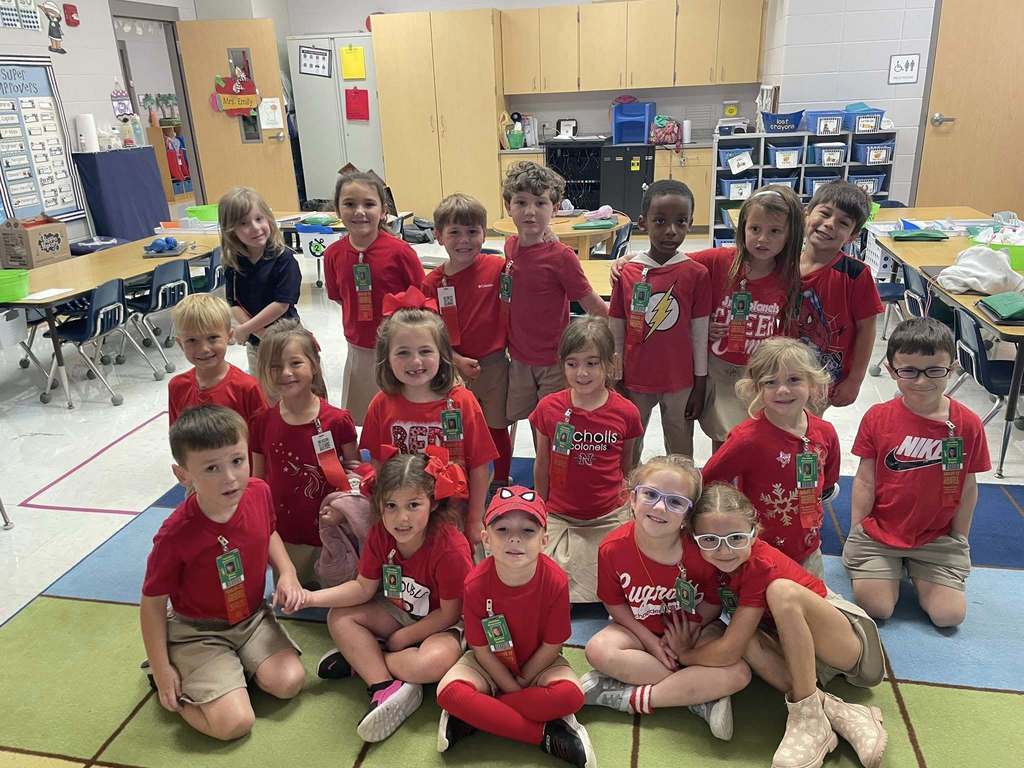 Group of children wearing bright, colorful shirts and khaki bottoms posing in front of a backdrop that says “RED RIBBON” with a large glittery red ribbon, celebrating Red Ribbon Week.