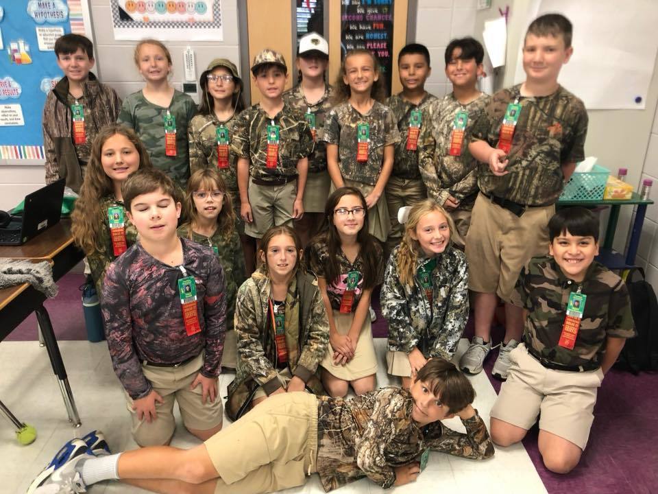 Group of children wearing bright, colorful shirts and khaki bottoms posing in front of a backdrop that says “RED RIBBON” with a large glittery red ribbon, celebrating Red Ribbon Week.