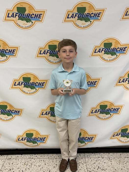 A student is standing in front of a backdrop featuring the Lafourche Parish Public Schools logo repeated across the background. The student is holding a trophy with both hands and is dressed in a light blue striped polo shirt, khaki pants, and brown shoes. The floor is speckled tile.