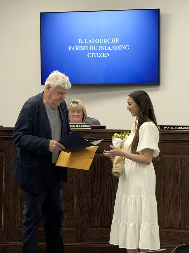 Madyson Cutrer, wearing a white dress and holding a bouquet of yellow roses, receives a certificate for the Lafourche Parish Outstanding Citizen Award from a man at a public meeting.