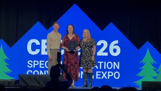 Three people stand on a stage in front of a large blue backdrop featuring a mountain silhouette and the text "CEC 2026 Special Education Convention & Expo." The person in the center, wearing a red floral dress, holds a plaque. They are flanked by a man in a tan shirt and grey trousers on the left, and a woman in a black and white floral wrap dress and black boots on the right.