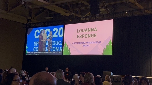 A wide shot of a large convention hall with an audience seated in the foreground. On a large elevated screen, the right side displays the name "LOUANNA ESPONGE" above the text "OUTSTANDING PARAEDUCATOR AWARD" against a light pink background with mountain and tree graphics. The left side of the screen shows a live video feed of the award presentation on stage.