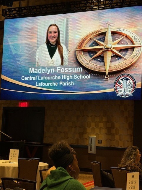 A photograph of a large projection screen at an event, displaying a recognition slide for a student. The slide features a portrait of a smiling young woman with long brown hair, identified as Madelyn Fossum from Central Lafourche High School, Lafourche Parish. To the right of her photo is a large, ornate golden compass rose design. The compass points are labeled with various educational program terms including "IDEA," "IREAP," and various "Title" designations (Title I, II, and III). In the bottom right corner of the slide is a circular logo for "LAFEPA" (Louisiana Association of Federal Program Administrators) featuring an image of the Louisiana State Capitol. The screen is situated in a dimly lit conference room with attendees seated at round tables in the foreground.