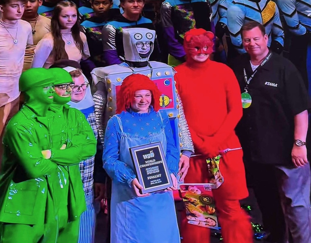A close-up photo of several students in vibrant costumes celebrating an achievement.  Central Figures In the Center: A student dressed as Raggedy Ann (blue patterned dress with a white apron and a bright red yarn wig) smiling and holding a black award plaque. The plaque reads: "WGI World Championships Percussion Scholastic Open Class FINALIST - Dayton, Ohio April 18, 2026."  To the Left: Two students dressed as green toy soldiers with their faces painted entirely green. Behind them, another student is partially visible in a blue and white striped outfit.  To the Right: A student in a solid red body suit with a red mask and a student dressed as a silver and grey robot.  Far Right: An adult male in a black polo shirt with a "TAMA" logo and a staff lanyard smiling at the group.  Background Other students in various performance costumes are visible in the background, including some in blue patterned athletic wear. The lighting is dramatic, typical of an indoor arena or stage setting.
