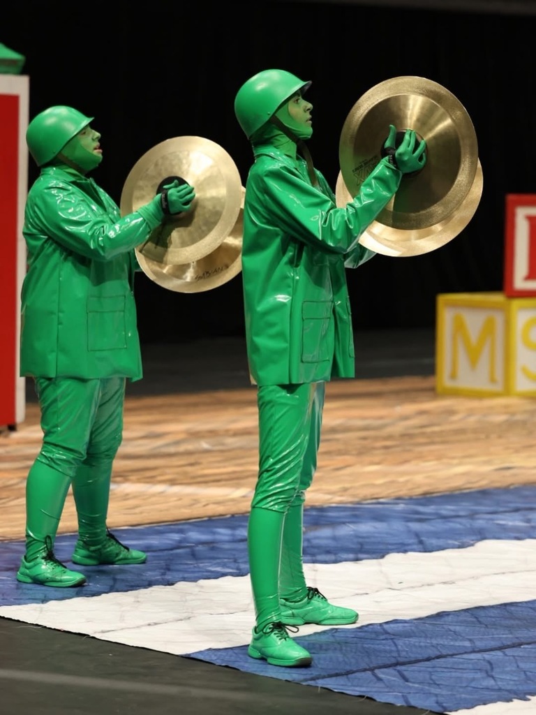 A dynamic photo of two students performing on a stage, dressed in elaborate, head-to-toe costumes that transform them into shiny green toy soldiers.  Central Figures The two performers stand on a stage mat, angled toward the right. Both wear a consistent monochromatic costume: a high-gloss green jacket and pants, green gloves, and a bright green helmet with their faces painted entirely green.  Left Performer: Holds a large pair of bronze-gold cymbals vertically in a crash position.  Right Performer: Holds a larger set of vertically stacked cymbals, looking forward as they hold the instrument in place.  Background Details The performance takes place on a wooden stage floor. In the background, out-of-focus yellow and red blocks with white letters (one showing 'M') suggest a whimsical, playroom-style setting. The lighting is focused and warm, typical of a performance arena.