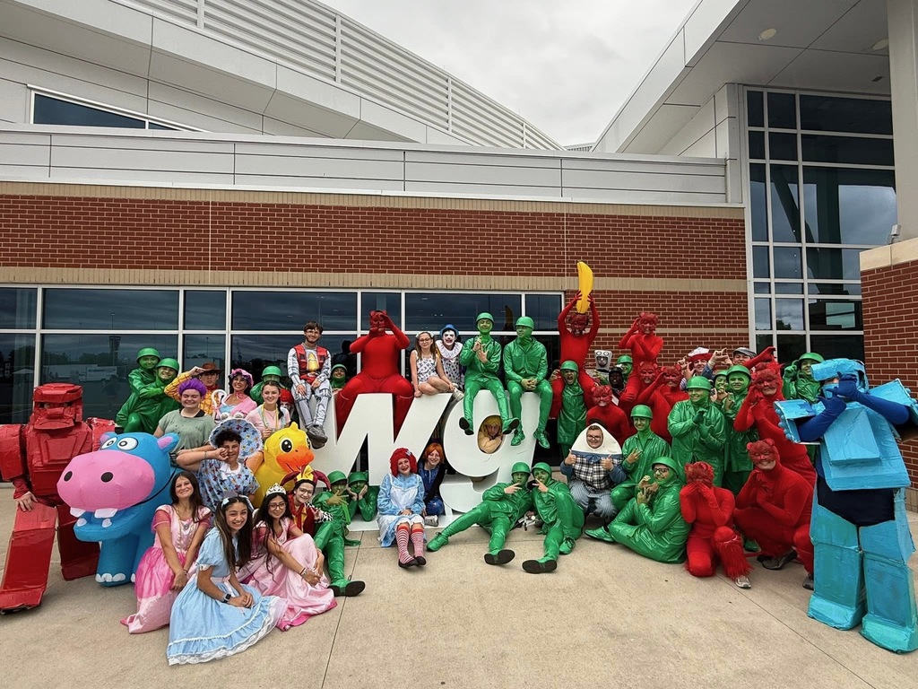 A group photo of dozens of students in vibrant, elaborate costumes posing in front of a modern brick and glass school building.  Group Description The students are gathered around a large, white decorative "W" and "G" sculpture on a concrete plaza. The costumes are diverse and colorful, including:  Monochromatic Groups: Multiple students dressed entirely in bright green as toy soldiers and others in solid red body suits.  Character Costumes: Individuals dressed as a pink hippopotamus, a yellow rubber duck, Raggedy Ann, princesses in pink and blue gowns, and a student in a red cardboard robot suit.  Whimsical Outfits: Students wearing inflatable suits, clown makeup, a banana costume, and a Humpty Dumpty-style egg outfit.  Background The background features the exterior of a high school with large windows reflecting the overcast sky and a contemporary white metal facade above the brickwork. The overall mood of the photo is festive and high-energy.