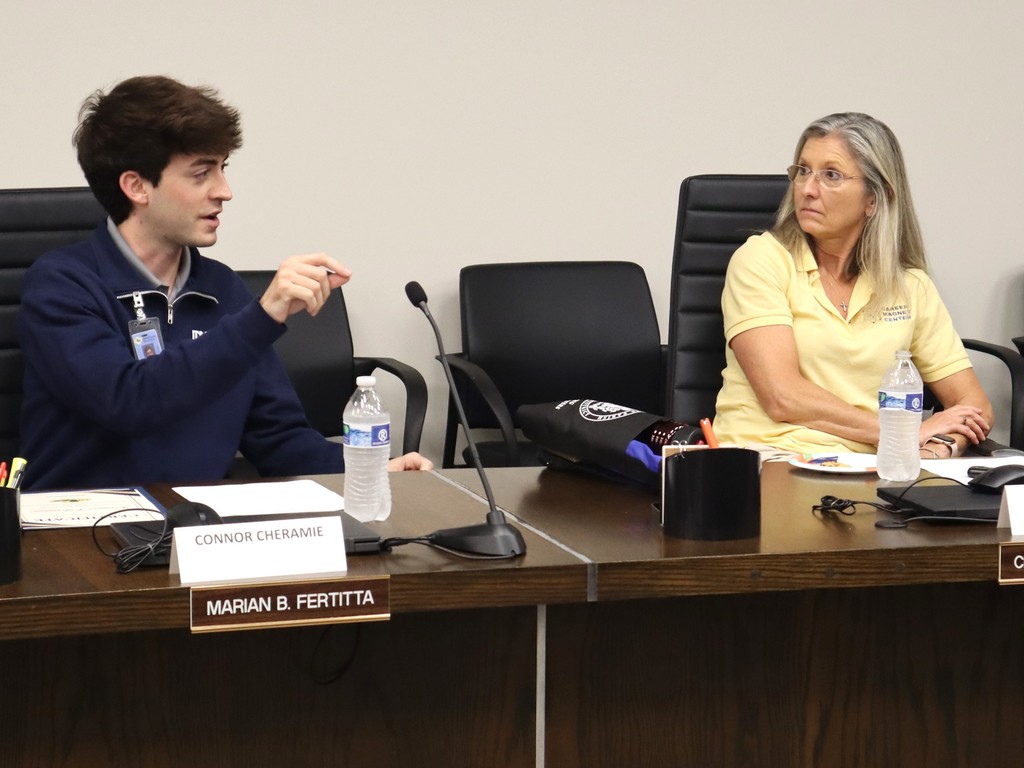 A candid photo capturing a focused exchange during a professional meeting at the Lafourche Parish School Board.  People & Action Left: A young man in a navy blue quarter-zip jacket is speaking and gesturing with a pen in his right hand. He is seated behind a nameplate that reads "CONNOR CHERAMIE" and a formal gold-on-black plate that reads "MARIAN B. FERTITTA."  Right: A woman in a yellow polo shirt with a "Career Magnet Center" logo is seated at the table, leaning forward slightly and listening intently to the speaker.  Setting & Details The Table: A dark wood conference table with microphones, bottles of water, a computer mouse, and a black padded bag with a white district logo.  The Background: A neutral-colored wall with several black mesh office chairs positioned behind the participants.  The image illustrates active engagement and dialogue between different generations within a district advisory or board setting.