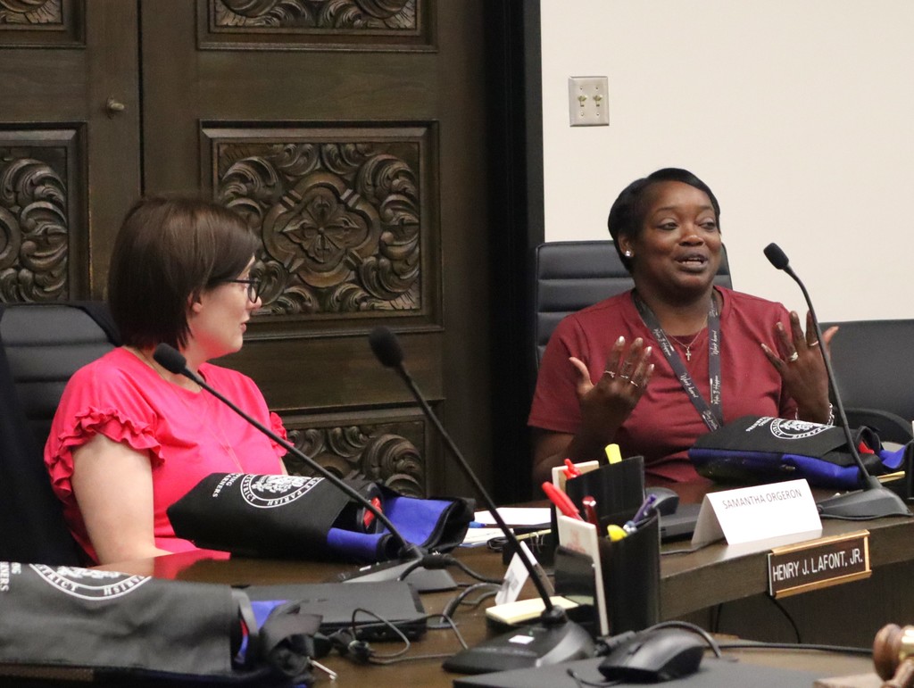 A candid photo of two women engaged in discussion during a professional meeting at the Lafourche Parish School Board.  People & Action Foreground Right: A woman in a maroon t-shirt with a lanyard is speaking expressively, using her hands to emphasize a point. She is seated behind a nameplate that reads "SAMANTHA ORGERON." * Foreground Left: A woman in a bright pink blouse with ruffled sleeves sits at the same table, looking toward the speaker and listening attentively.  Setting & Details The Table: A dark wood conference table equipped with several gooseneck microphones, a computer mouse, and office supplies. Large black and blue padded bags with a circular white logo are resting on the table in front of each participant.  The Background: Behind the women is a set of large, dark wood double doors featuring intricate, ornate floral carvings.  Nameplates: In addition to Samantha Orgeron's nameplate, a formal gold-on-black nameplate at the edge of the table reads "HENRY J. LAFONT, JR."  The image captures a moment of active collaboration and dialogue within a formal district meeting setting.