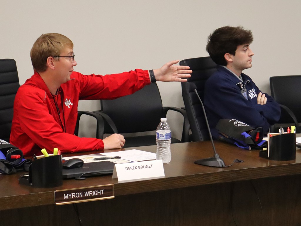 A candid photo of two young men participating in a professional meeting at the Lafourche Parish School Board.  People & Action Left: A young man in a red jacket is gesturing with an open hand toward the center of the room while speaking. He is seated behind a nameplate that reads "DEREK BRUNET" and a formal gold-on-black plate that reads "MYRON WRIGHT."  Right: Another young man in a navy blue quarter-zip jacket sits with his arms crossed, listening attentively to the discussion.  Setting & Details The Table: A dark wood conference table featuring a computer mouse, a bottle of water, a microphone, and a cup of pens/highlighters.  The Background: A neutral-colored office wall with extra black mesh office chairs lined up behind the participants.  The image captures an active moment of student or employee participation in a formal advisory council setting.