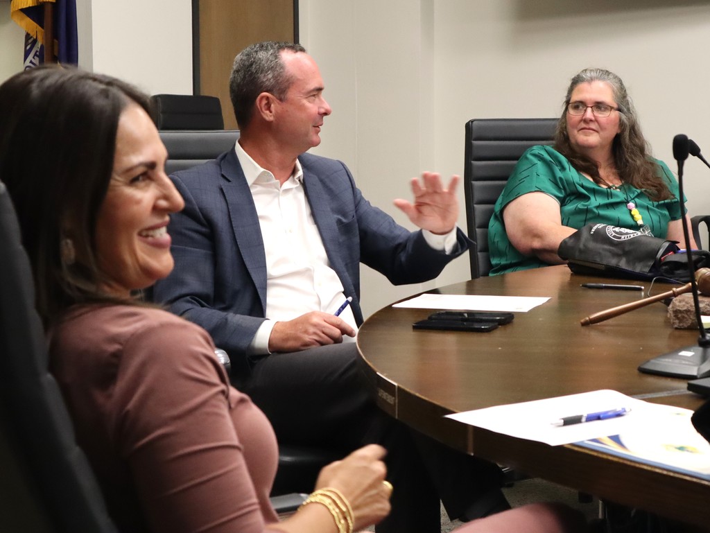 A candid photo capturing a moment of lighthearted discussion during a professional meeting at the Lafourche Parish School Board.  People & Action Foreground Left: A woman in a mauve-colored dress is smiling broadly, shown in profile as she looks toward the center of the table.  Center: A man in a blue blazer and white dress shirt (Superintendent Martin) is speaking and gesturing with an open hand while looking toward the right. He is holding a blue pen.  Background Right: A woman in a green patterned blouse is seated at the table, listening attentively with a slight smile.  Setting & Details The Table: The group is seated at a large, dark wood oval conference table. Visible on the table are white documents, blue pens, a smartphone, and a wooden gavel resting on a stone block.  The Atmosphere: The lighting is bright and professional, and the expressions suggest a positive, collaborative environment.  This image highlights the human element of district leadership and advisory council meetings.