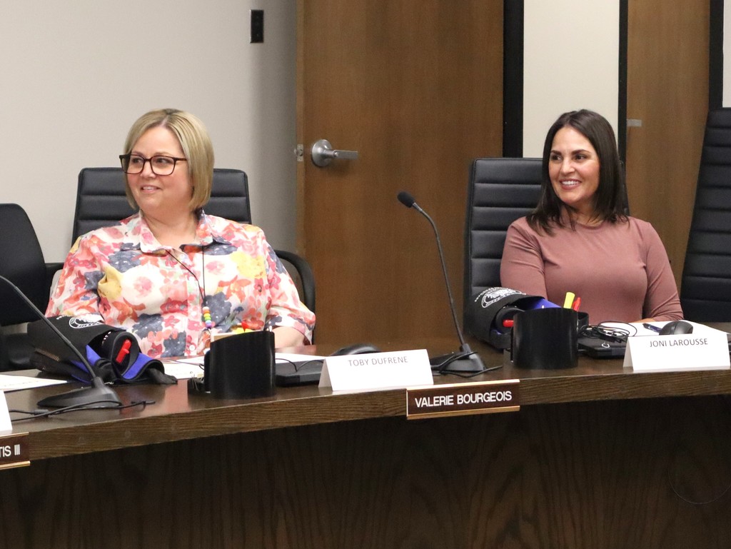 A candid photo of two women participating in a professional meeting at the Lafourche Parish School Board.  People & Action Foreground Left: A woman with blonde hair and glasses, wearing a colorful floral blouse, sits at the table and looks toward the center of the room with a smile.  Foreground Right: A woman with dark hair, wearing a mauve-colored top, is also seated at the table, smiling and looking in the same direction.  Setting & Details The Table: Both women are seated at a dark wood conference table equipped with gooseneck microphones, a computer mouse, and a cup of pens.  Nameplates: White triangular nameplates on the table identify the participants as Toby Dufrene and Joni Larousse. A formal gold-on-black nameplate at the edge of the table reads "VALERIE BOURGEOIS."  Background: A neutral-colored wall and a wooden door with a silver handle are visible behind the participants.  The image captures a friendly and collaborative atmosphere during a formal district meeting.