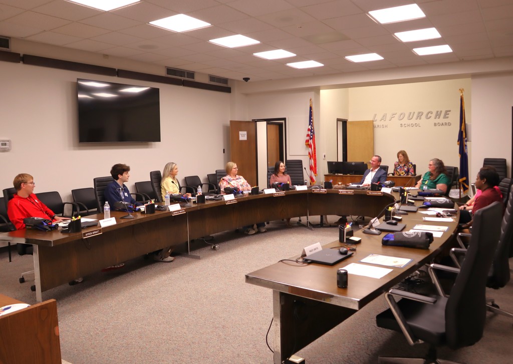 A wide-angle photo of a professional meeting taking place in a large boardroom at the Lafourche Parish School Board.  Setting & Atmosphere The room is bright with fluorescent ceiling lights and features a neutral grey carpet. A large, horseshoe-shaped dark wood conference table dominates the space, equipped with microphones, laptops, and black-and-blue gear bags at several seats. On the left wall, a large flat-screen television is mounted, though it appears to be turned off.  Participants Ten individuals are seated around the table, including:  Left side: Two young men (one in a red jacket, one in a navy quarter-zip) and a woman in a yellow polo.  Center/Head of Table: Superintendent Martin (in a blue blazer) is seated near the center, with several other women in professional attire (floral, mauve, and green tops) positioned around the curve.  Right side: A woman in a maroon t-shirt is seated facing the rest of the group.  Background Details In the back right corner, the words "LAFOURCHE PARISH SCHOOL BOARD" are mounted on the wall in silver lettering.  An American flag and a blue state or district flag stand on poles in the background.  A wooden door in the center background stands slightly ajar.  The image captures a formal yet collaborative meeting environment, likely a session of the Superintendent’s Advisory Council.