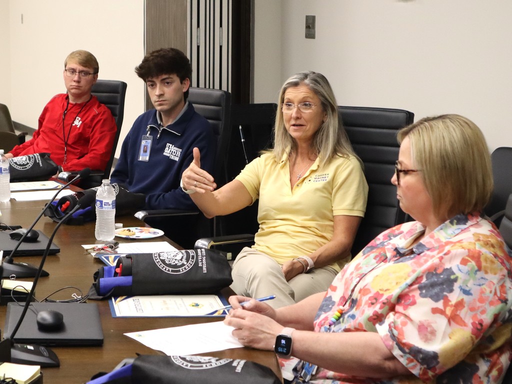 A candid photo capturing a discussion during a professional meeting at the Lafourche Parish School Board.  People & Action Center: A woman in a yellow polo shirt with a "Career Magnet Center" logo is speaking and gesturing with her hand to make a point.  Background Left: Two young men are seated at the table, listening intently to the speaker. One wears a red jacket and the other wears a navy blue quarter-zip.  Foreground Right: A woman in a floral patterned blouse is seated at the table, looking down at her notes and holding a blue pen. She is wearing a black smartwatch on her left wrist.  Setting & Details The Table: A dark wood conference table equipped with gooseneck microphones, computer mice, and bottles of water. Several black and blue padded bags with the district logo are placed in front of the participants.  Documents: A certificate and other meeting papers are visible on the table surface.  Atmosphere: The setting is a formal boardroom with neutral-colored walls and black leather office chairs.  The image illustrates a collaborative dialogue between  staff during an advisory council  session.