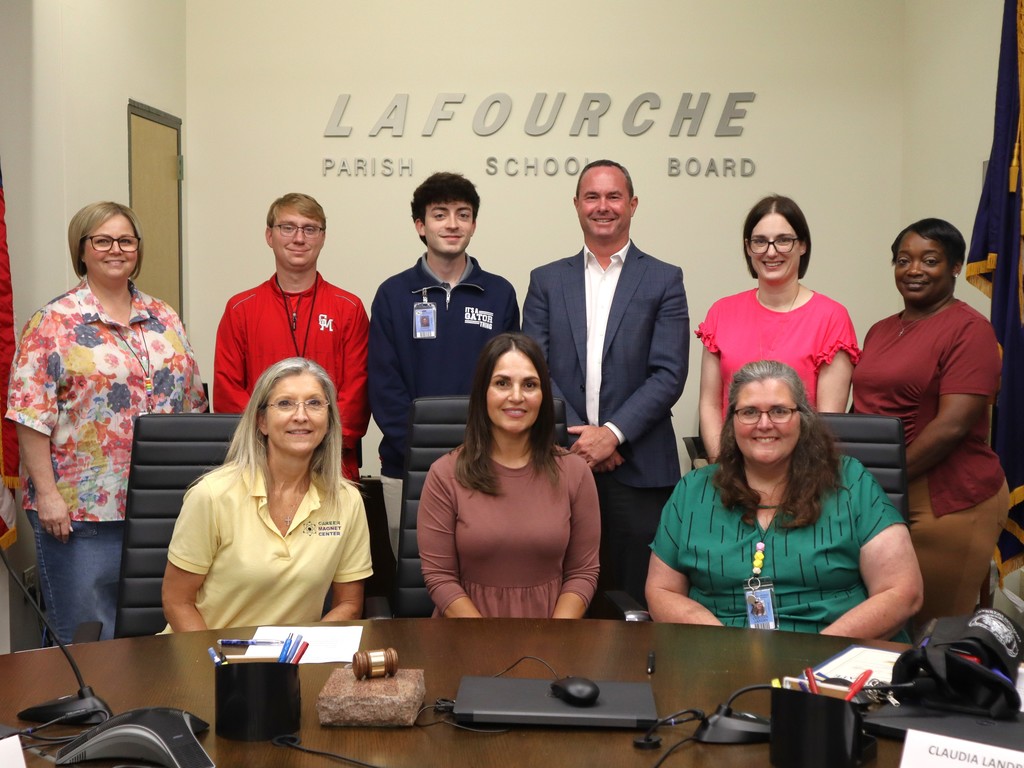 A group photo of nine people gathered in a professional meeting room at the Lafourche Parish School Board office.  People & Setting The group is posed around a large, dark wood oval conference table.  Front Row: Three women are seated at the table. The woman on the left wears a yellow polo shirt with a "Career Magnet Center" logo, the woman in the center wears a mauve-colored dress, and the woman on the right wears a green patterned blouse.  Back Row: Six people are standing behind the seated group. From left to right: a woman in a floral blouse, a young man in a red jacket, a young man in a navy quarter-zip, a man in a blue blazer and white shirt (Superintendent Martin), a woman in a bright pink top, and a woman in a maroon shirt.  Room Details Background: A neutral-colored wall features large, raised silver lettering that reads "LAFOURCHE PARISH SCHOOL BOARD." * Table Items: On the table are microphones, a gavel resting on a stone block, pens in a cup, and some folders.  Flags: To the far right, a portion of a blue flag with gold fringe is visible.  The overall atmosphere is professional and collaborative, typical of a board or committee meeting.