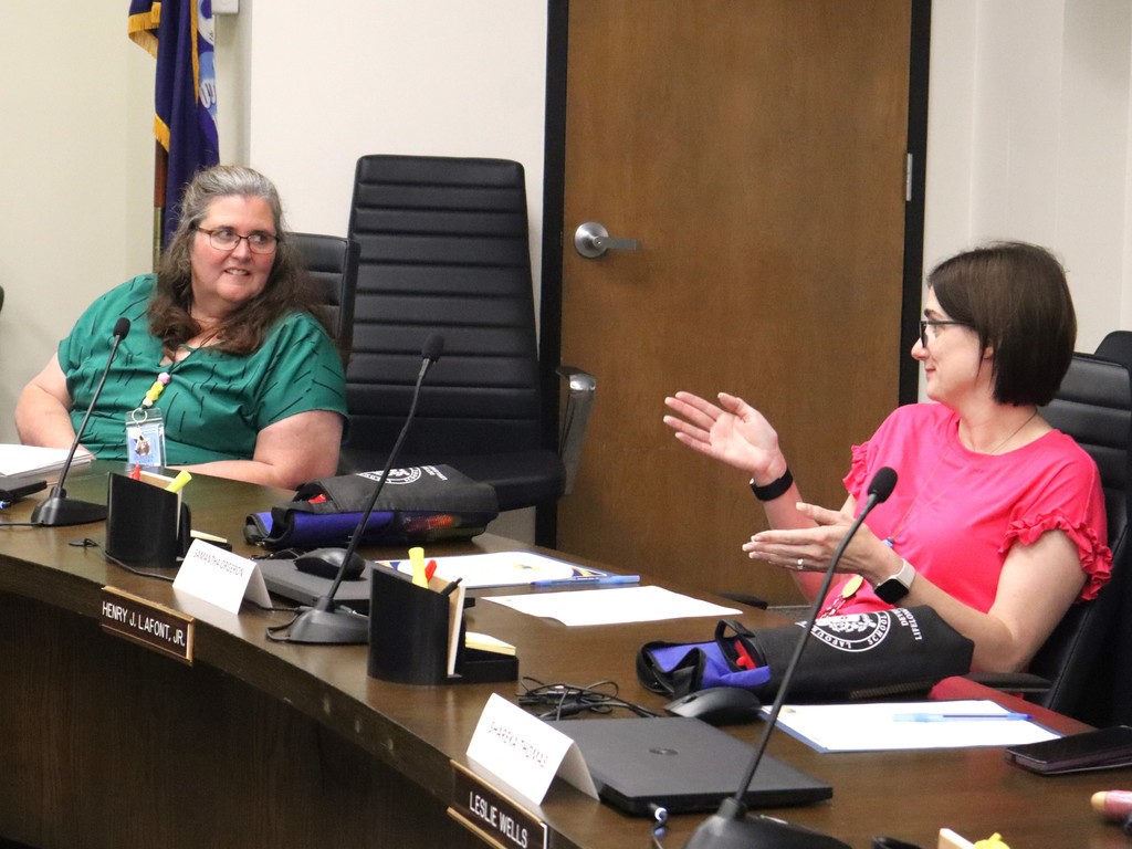 A candid photo of two women engaged in discussion during a meeting at the Lafourche Parish School Board.  People & Action Foreground Right: A woman with dark hair and glasses, wearing a bright pink blouse with ruffled sleeves, is speaking and gesturing with both hands open. She sits behind a nameplate that reads "SHAREKA THOMAS."  Foreground Left: A woman with long brown hair and glasses, wearing a green patterned top, sits at the same table and looks toward the speaker with an attentive smile. She is seated behind a nameplate that reads "SAMANTHA ORGERON."  Setting & Details The Table: A dark wood conference table equipped with several gooseneck microphones, computer mice, and cups of pens/highlighters. Two black and blue padded gear bags with a circular district logo are placed on the table.  Background: A neutral-colored wall and a wooden door are visible. A formal gold-on-black nameplate at the edge of the table reads "LESLIE WELLS," and another partially visible nameplate reads "HENRY J. LAFONT, JR."  Flags: A portion of a blue flag with gold fringe is visible in the upper left corner.  The image captures a moment of active listening and collaborative dialogue during a formal meeting session.
