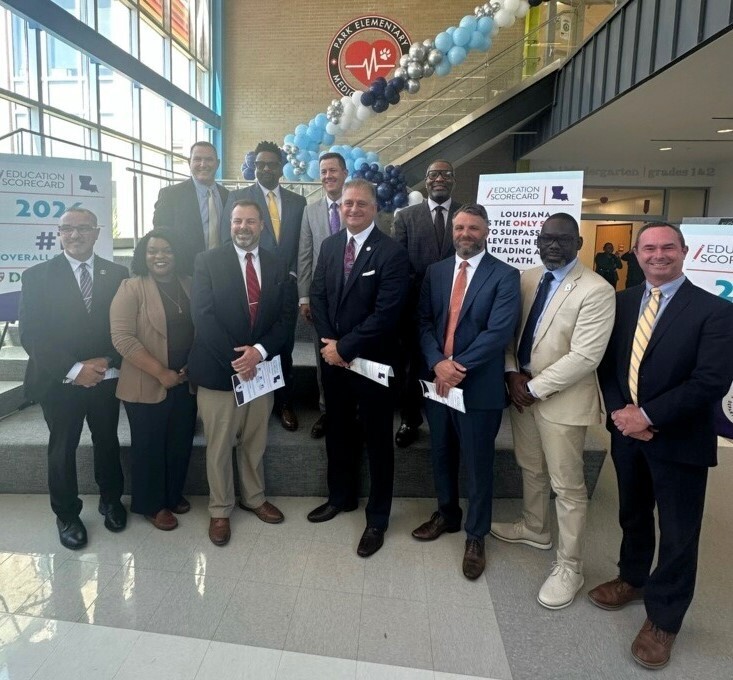 A group of education officials and school leaders posing for a photo on a set of stairs inside Park Elementary Medical Academy. The group is dressed in professional attire and stands in front of "Education Scorecard" posters. A blue, white, and silver balloon arch curves behind them toward the school’s logo on the wall. Large windows to the left fill the modern lobby with natural light. Lafourche Parish School District Superintendent Jarod Martin is on the right.