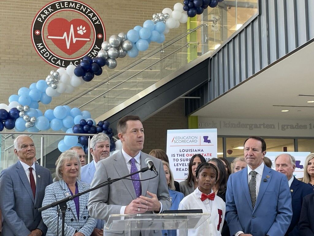 Louisiana State Superintendent of Education Cade Brumley speaks at a podium during a press conference at Park Elementary Medical Academy. He is surrounded by a group of officials and a young student in a red bowtie. In the background, a large "Education Scorecard" sign is visible alongside a blue, white, and silver balloon arch. The school's circular logo, featuring a heart and pulse line, is mounted on the brick wall behind the group.