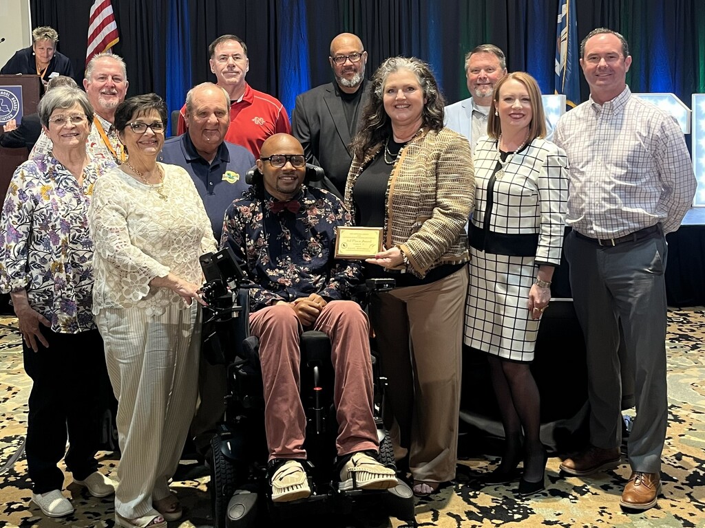 A group photo of eleven people gathered on a stage for an awards presentation, standing in front of dark blue and green curtains. In the center foreground, a man seated in a motorized wheelchair wears a dark floral shirt, a red bow tie, pink pants, and glasses. He is holding a small gold plaque alongside a woman standing to his right who has long, dark wavy hair with grey streaks, wearing a patterned brown jacket and tan pants. Surrounding them are other attendees smiling for the camera: To the left of the wheelchair, a woman in a white lace blouse and cream pants stands next to a woman in a purple and yellow floral shirt. To the right of the woman in the brown jacket, a woman in a black-and-white grid-pattern skirt suit stands next to a man in a plaid button-down shirt and grey slacks. Several other individuals stand in the background row, including a man in a red polo shirt, a man in a black blazer, and a man in a light blue blazer. An American flag and a state flag are partially visible in the background.