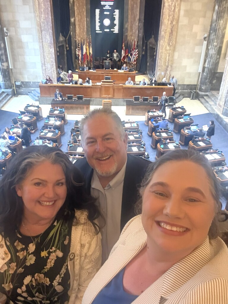 A high-angle selfie of three smiling people in the foreground, looking up at the camera from an elevated gallery overlooking a large legislative chamber. In the foreground from left to right: A woman with long, dark wavy hair with grey streaks, wearing a dark floral top and a white cardigan. A man with short grey hair and a goatee, wearing a grey button-down shirt and a dark blazer. A woman with lighter hair tied back, wearing a striped blazer over a blue top, holding the camera. In the background, the ornate legislative floor features rows of dark wooden desks arranged in a semi-circle on a blue carpeted floor, with several people seated or standing. At the far end of the room is a large wooden dais with a speaker's podium, multiple flags flanking the stage, and an electronic voting board mounted high on the wall.