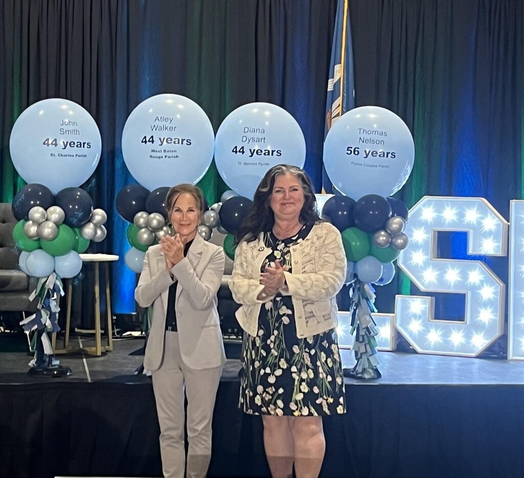 An image of an awards ceremony stage featuring two women standing and clapping in the foreground, with four large light-blue milestone balloons arranged behind them. The woman on the left has blonde hair tied back, wearing a light-grey pantsuit over a dark top. The woman on the right has long, dark wavy hair with grey streaks, wearing a floral dress and a cream-colored cardigan. Behind them, four identical balloon bouquets feature large, round light-blue top balloons, each printed with a person's name and years of service, surrounded by smaller dark blue, green, and silver balloons. From left to right, the text on the large balloons reads: "John Smith, 44 years, St. Charles Parish" "Alley Walker, 44 years, West Baton Rouge Parish" "Diana Dysart, 44 years, St. Bernard Parish" "Thomas Nelson, 56 years, Pointe Coupee Parish" To the far right, a large marquee letter "S" lit with bright bulbs is partially visible. The background consists of dark blue and green curtains.