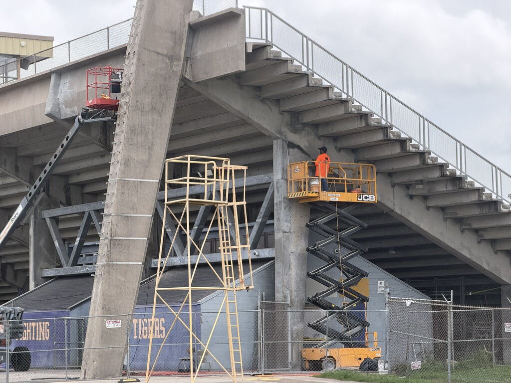 An outdoor shot showing maintenance and concrete repair work underneath a large stadium grandstand structure. In the center right, a worker wearing a bright orange high-visibility shirt and a hard hat stands in the yellow basket of an elevated JCB scissor lift. They are using a tool to patch or work on a thick concrete support pillar beneath the tiered stadium seating. To the far left, a red boom lift basket is extended high up near a slanted, vertical concrete support column. In the foreground, a tall, yellow mobile scaffolding tower stands on the concrete ground. Behind a chain-link security fence at the base of the grandstands, a purple building structure is visible with the words "FIGHTING TIGERS" painted on it in large yellow block letters. The sky overhead is overcast and cloudy.