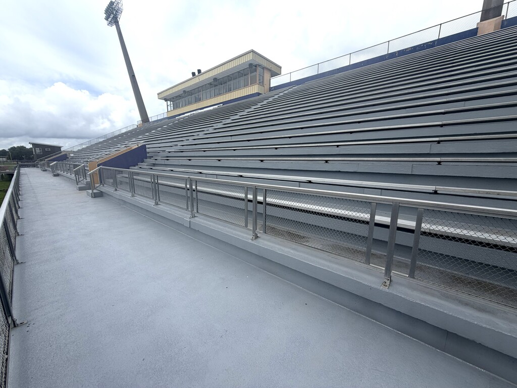 A wide-angle, low-angle shot of a large, empty outdoor stadium grandstand under a cloudy sky. The perspective looks up and across a wide, smooth grey concrete walkway or concourse area in the foreground. To the right, tiers of grey concrete bleacher seating with long metal benches rise steeply toward the top of the stadium. A stainless steel safety railing with chain-link mesh panels separates the walkway from the seating area. In the upper-left background, a two-story press box with large glass windows sits at the top of the grandstands, next to a tall stadium light pole.