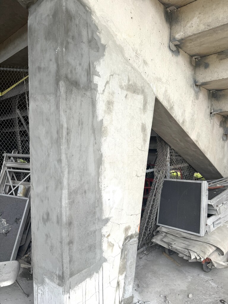 A close-up shot of a thick, vertical concrete support pillar beneath a stadium showing visible repair work. The left side and upper edge of the pillar have been patched with fresh, smooth, dark grey concrete, contrasting with the aged, weathered off-white concrete of the original structure. Fine surface cracks are visible on the older, unpatched sections of the column. In the background behind the pillar, a cluttered under-tier storage area is visible through a chain-link fence. The area contains stacked metal scaffolding pieces, large rectangular black road cases or panels, and folded tarps resting on a wheeled cart.