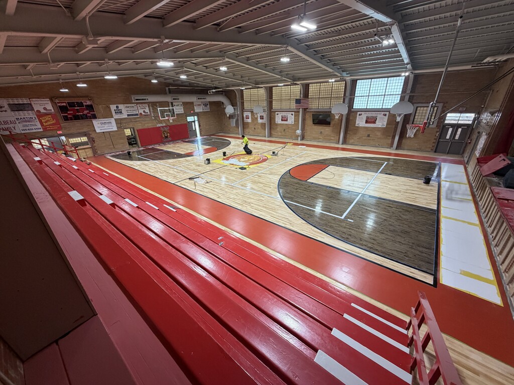 A high-angle, wide-view photograph looking down from the top of bright red bleachers onto an indoor basketball gymnasium undergoing floor maintenance.  The basketball court features polished light-wood flooring with dark-stained wood sections inside the three-point arcs, red painted keys, and a red perimeter border. At the center circle, a circular logo features a lion's head with the words "GOLDEN MEADOW LIONS". A worker in a high-visibility neon yellow shirt is operating a floor buffer near the center circle, surrounded by scattered maintenance buckets and equipment.  The gym has brick walls lined with local business sponsor banners, championship signs, a digital scoreboard on the left, and an American flag hanging on the far wall beneath a row of high windows. Basketball hoops with glass backboards are positioned at each end of the court.